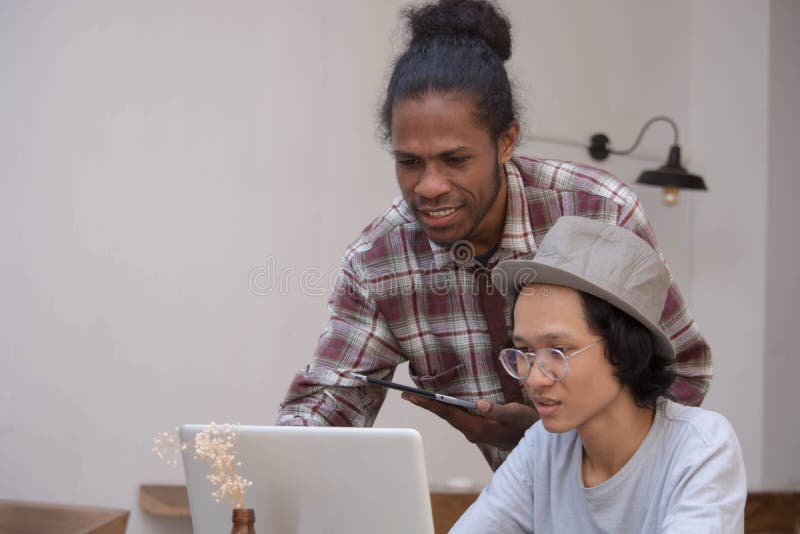 两年轻创造性的人与的膝上型计算机谈论和片剂、年轻工作亚洲人和的黑人与片剂和膝上型计算机在咖啡馆,巴布亚 库存图片 两年轻创造性的人与的膝上型计算机谈论和片剂、年轻工作亚洲人和的黑人与片剂和膝上型计算机在咖啡馆,巴布亚 库存图片